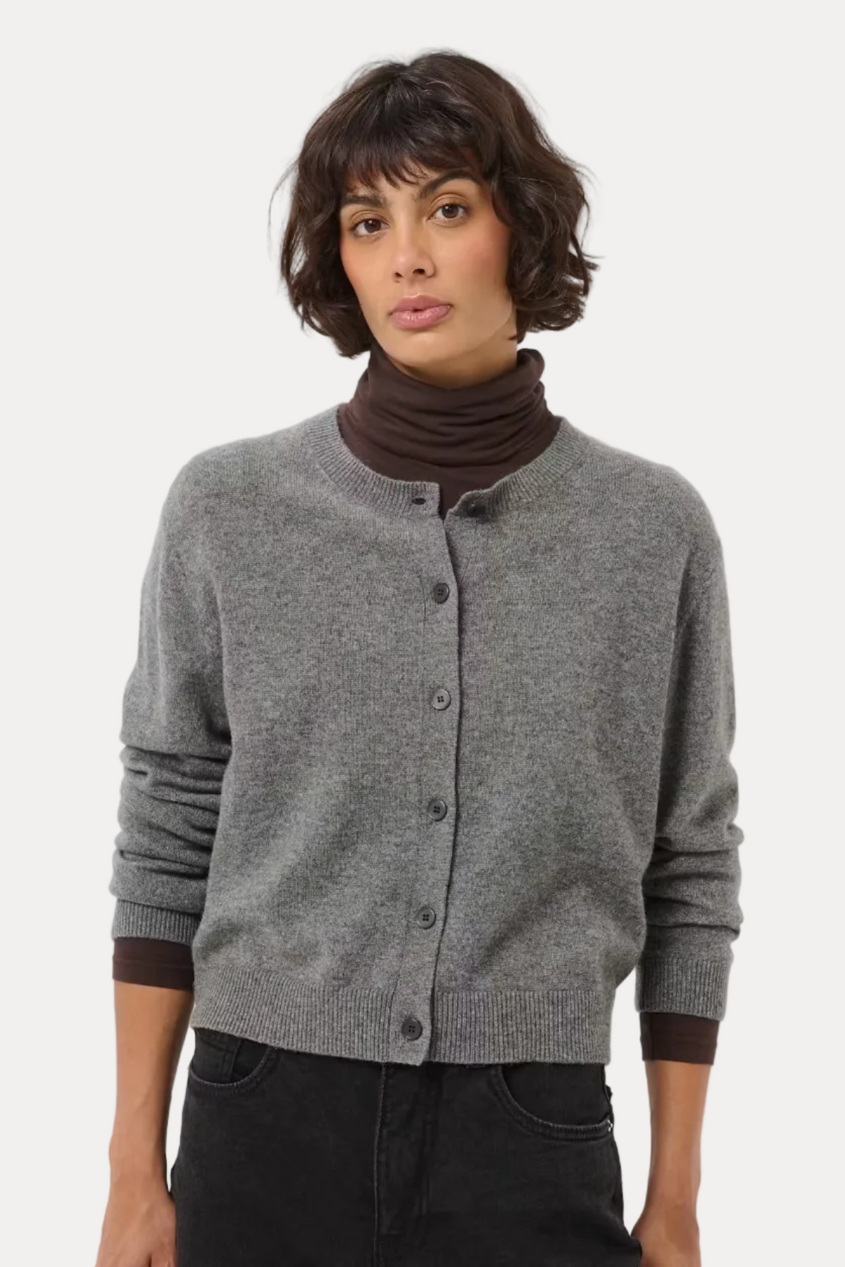 A model wearing a gray button-up cardigan over a dark brown ribbed turtleneck, paired with black pants, against a light gray studio background.