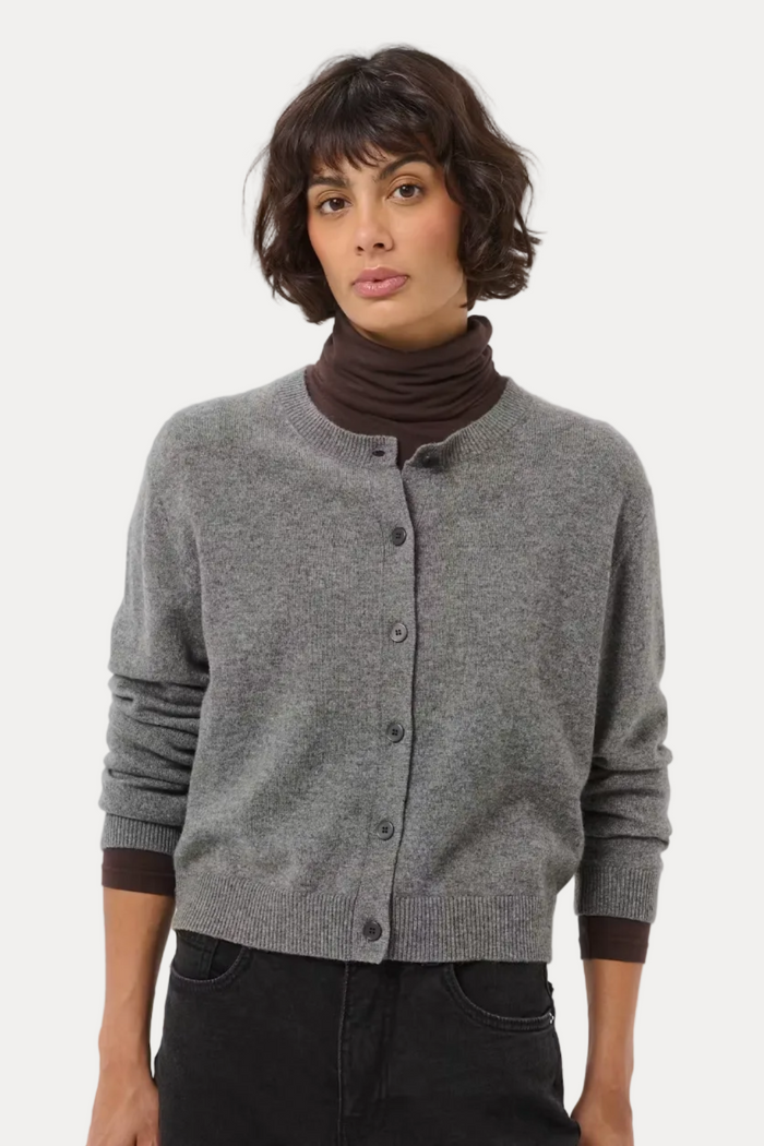A model wearing a gray button-up cardigan over a dark brown ribbed turtleneck, paired with black pants, against a light gray studio background.