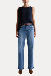 A model wearing a sleeveless dark navy top, blue wide-leg jeans, and brown suede boots, standing against a plain light gray studio background.