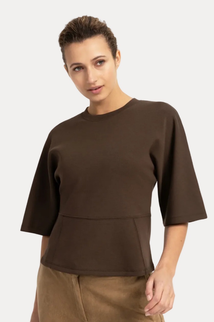 A woman modeling a dark brown short-sleeve knit top with a structured, slightly cropped silhouette and a peplum-like seam at the waist, paired with brown pants against a plain white studio background.