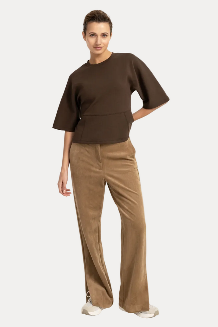 A model stands against a plain white studio background wearing a dark brown short-sleeve cropped top and tan wide-leg pants with white sneakers.