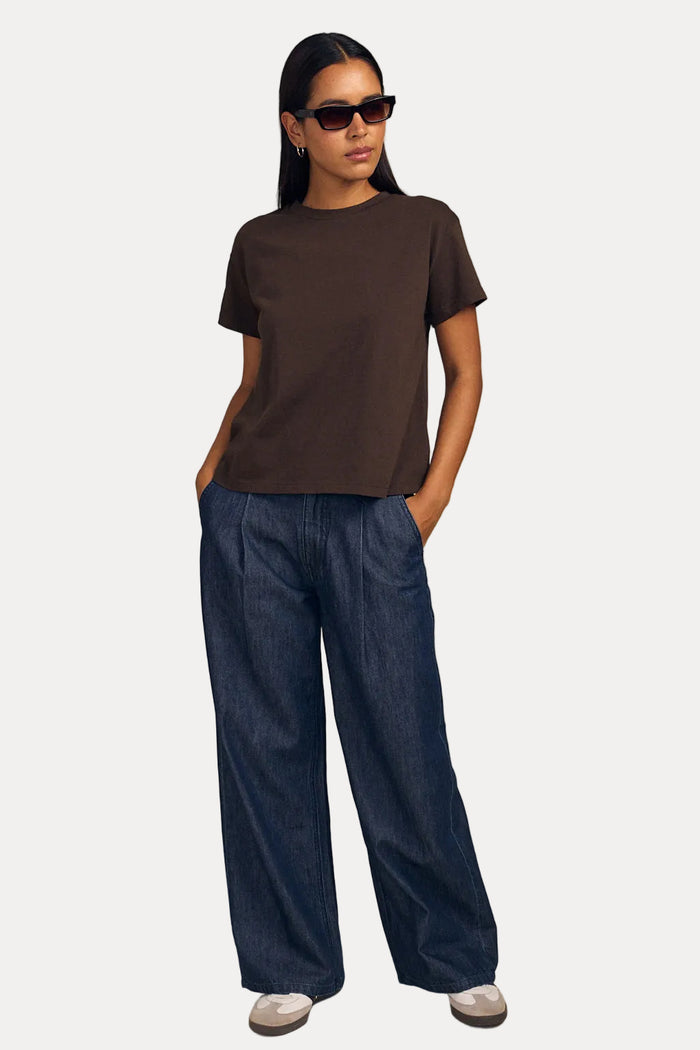 A woman modeling a dark brown short-sleeve crewneck t-shirt and wide-leg dark blue jeans against a solid beige studio background.