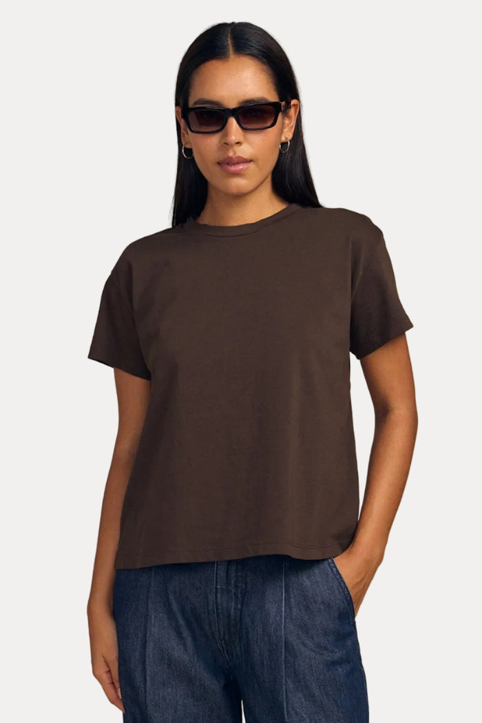A woman modeling a dark brown short-sleeve crewneck t-shirt and wide-leg blue jeans against a solid beige background.