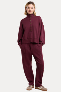 A model wearing a matching burgundy two-piece lounge set consisting of a long-sleeve button-up top and relaxed-fit pants, paired with beige sneakers, against a solid beige background.