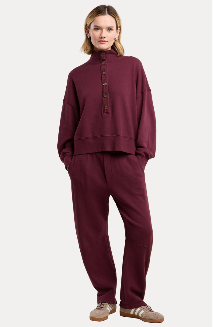 A model wearing a matching burgundy two-piece lounge set consisting of a long-sleeve button-up top and relaxed-fit pants, paired with beige sneakers, against a solid beige background.