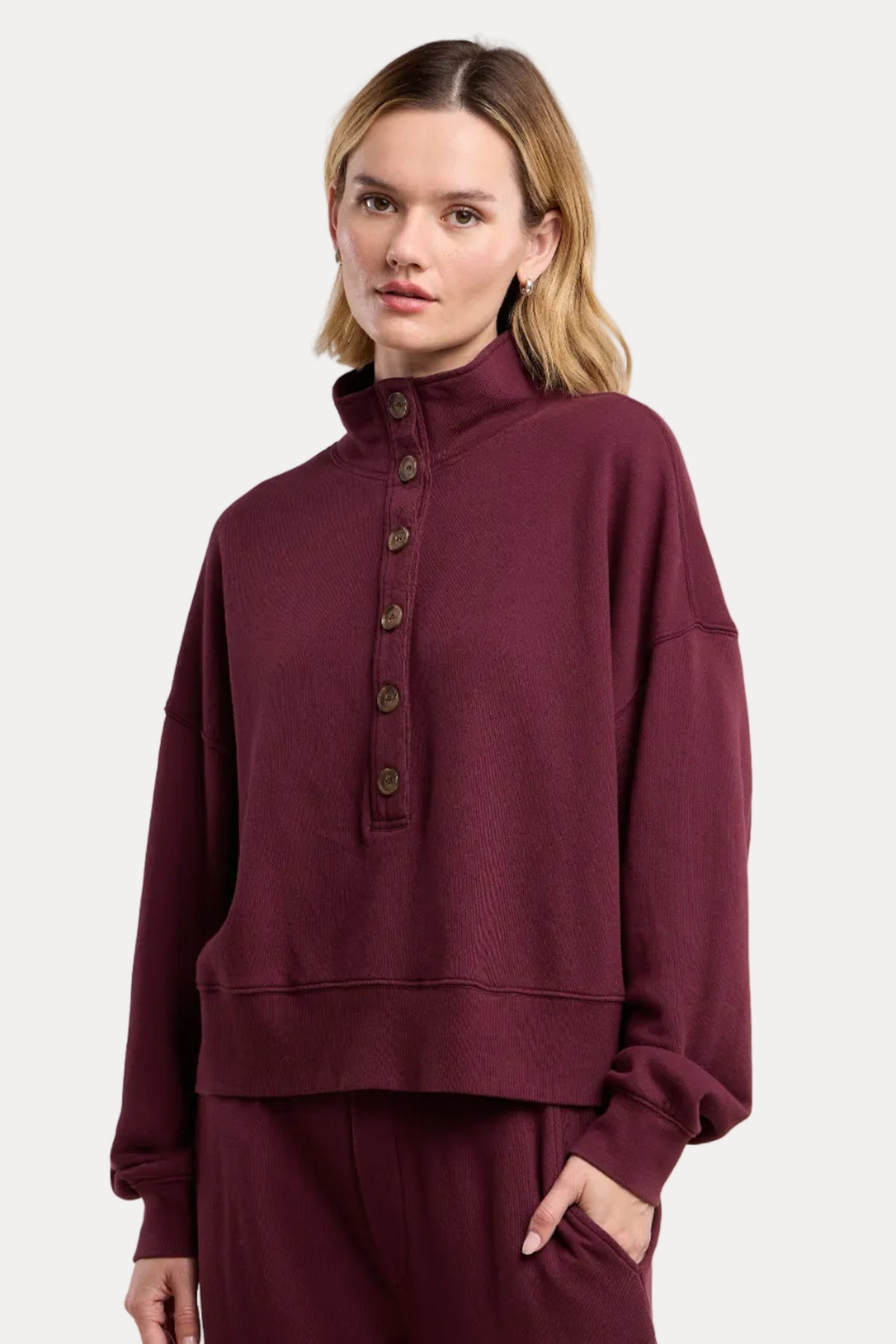 A woman modeling a matching burgundy lounge set, featuring a cropped long-sleeve sweatshirt with a high mock neck and button placket, paired with matching sweatpants, against a solid beige background.