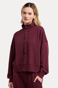 A woman modeling a matching burgundy lounge set, featuring a cropped long-sleeve sweatshirt with a high mock neck and button placket, paired with matching sweatpants, against a solid beige background.