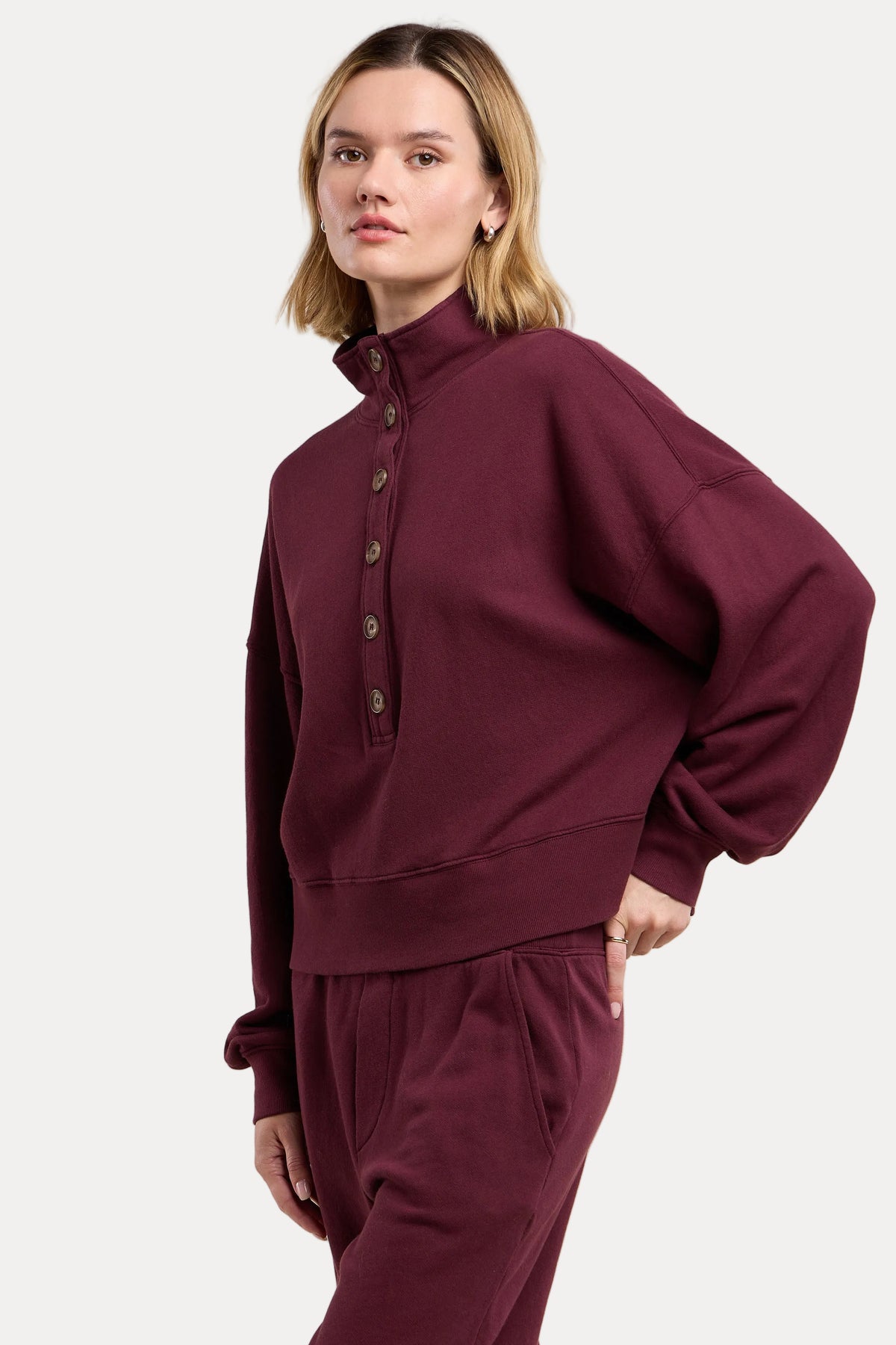 A woman modeling a matching burgundy sweatshirt and sweatpants set against a solid beige background.