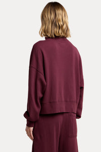 A model shown from the back wearing a matching burgundy crewneck sweatshirt and sweatpants set against a solid beige background.