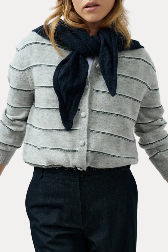 A woman wearing a light gray knit cardigan with thin dark horizontal stripes, a dark navy scarf tied around the neck, and dark trousers, shown against a plain light gray studio background.