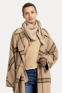 A woman modeling a long beige plaid coat over a beige sweater, blue jeans, and beige sneakers, standing against a plain white studio background.