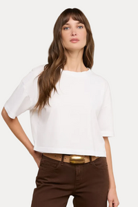 A woman modeling a white short-sleeve cropped t-shirt with brown pants and a gold-belt, against a solid off-white background.