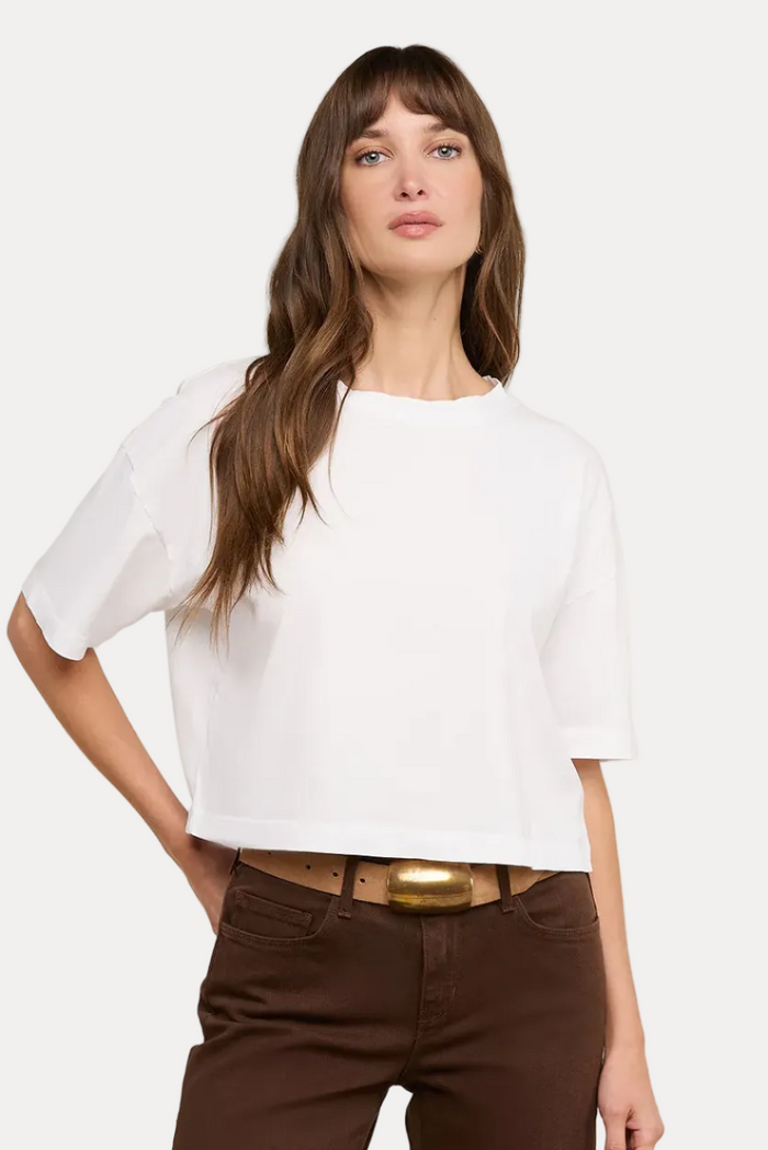 A woman modeling a white short-sleeve cropped t-shirt with brown pants and a gold-belt, against a solid off-white background.