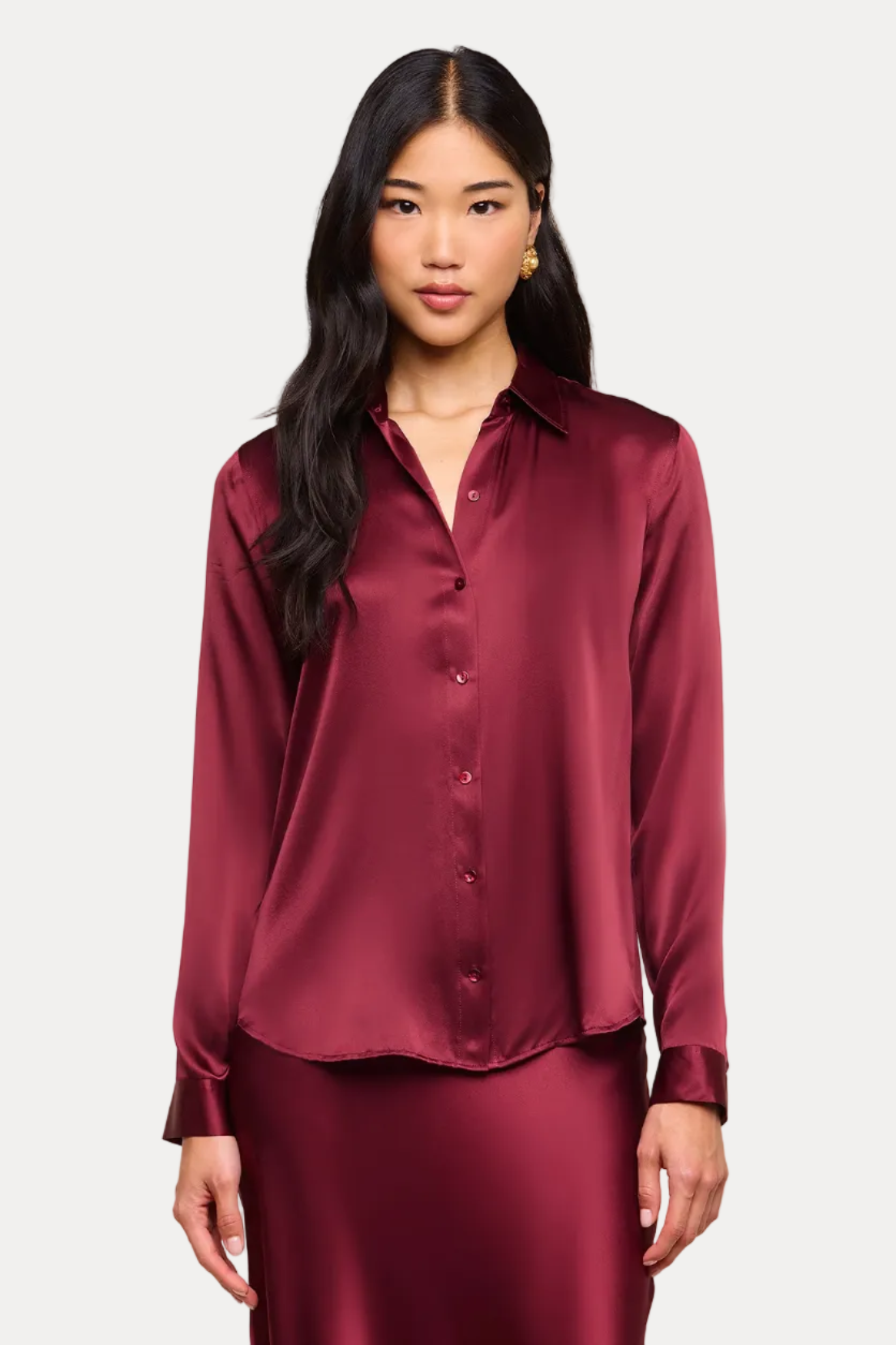 A woman modeling a matching burgundy satin button-up shirt and long skirt set against a solid beige studio background.