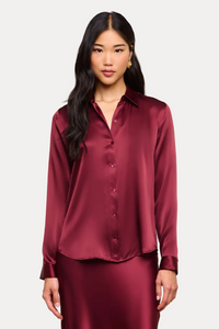 A woman modeling a matching burgundy satin button-up shirt and long skirt set against a solid beige studio background.