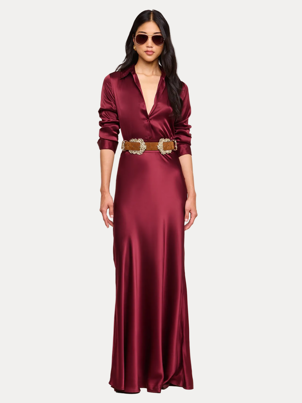 A woman modeling a floor-length satin maxi dress in deep burgundy with a wide brown belt featuring a large decorative buckle, set against a solid beige studio background.