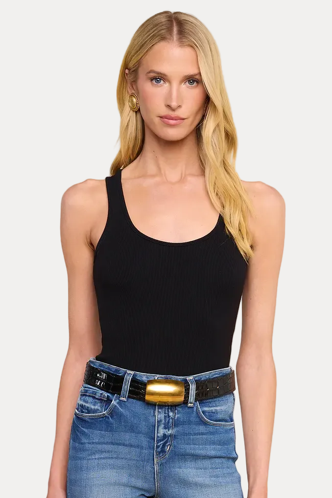 A blonde model wearing a black scoop-neck tank top and blue jeans with a black belt featuring a prominent gold buckle, set against a solid beige background.