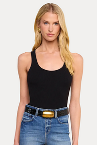A blonde model wearing a black scoop-neck tank top and blue jeans with a black belt featuring a prominent gold buckle, set against a solid beige background.