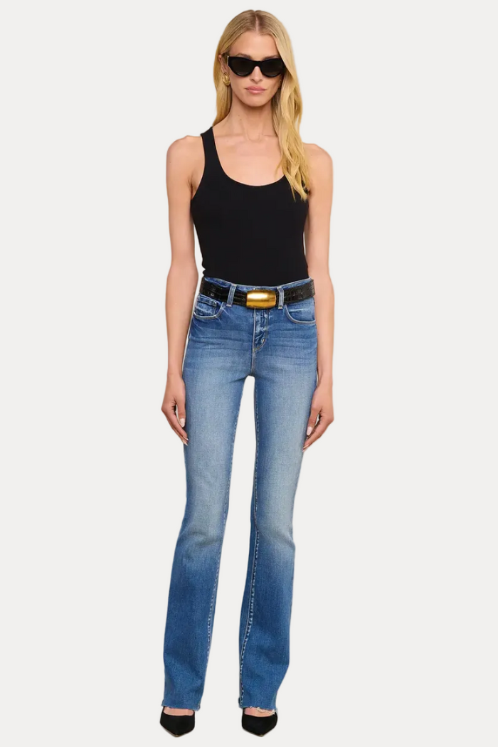 A woman modeling a pair of medium-wash blue jeans with a black belt featuring a gold buckle, styled with a black tank top and black sunglasses against a solid beige background.