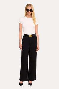 A woman modeling a white short-sleeve crewneck top and black wide-leg trousers with a gold belt, styled with black pointed-toe heels and sunglasses, set against a solid off-white studio background.