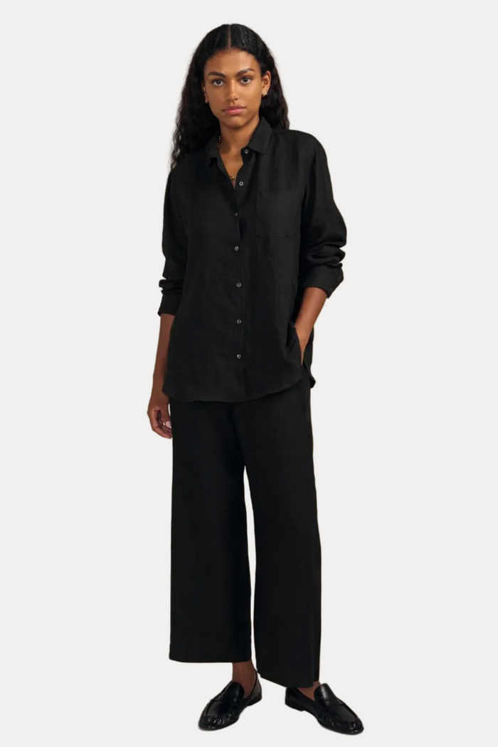 A model stands against a solid beige studio background wearing a black linen button-up shirt, matching black linen wide-leg trousers, and black leather loafers.
