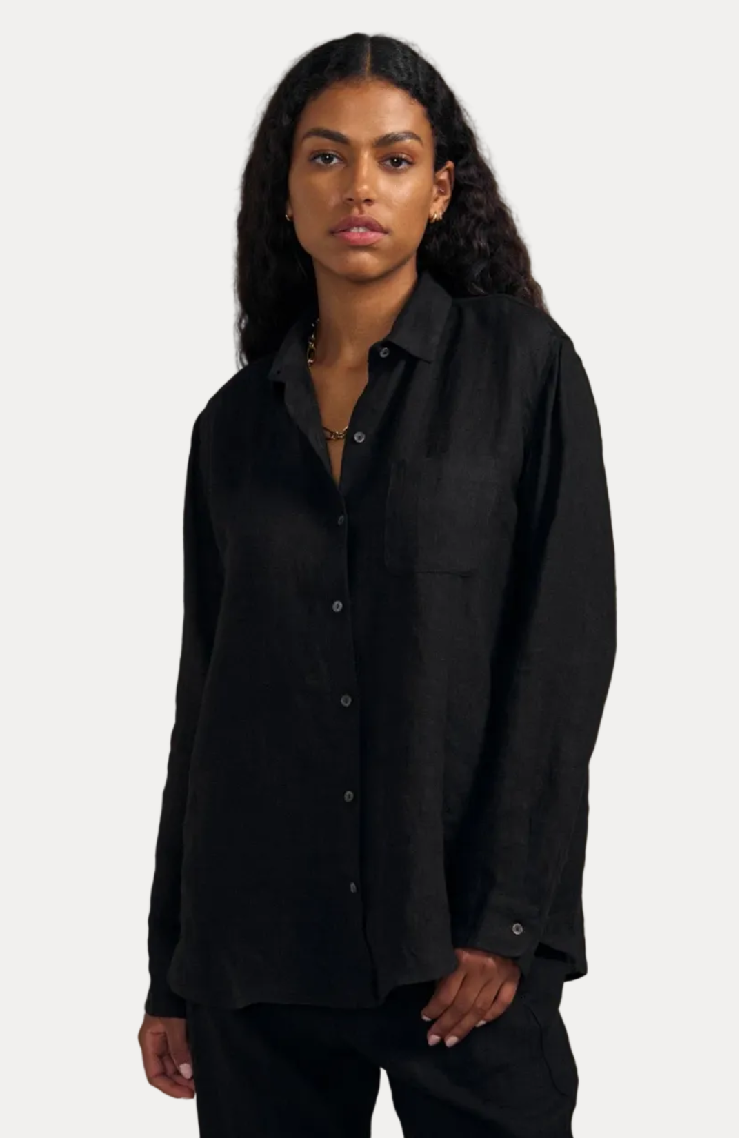A woman modeling a black long-sleeve linen button-up shirt with a chest pocket, paired with black pants, against a solid beige background.