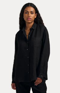 A woman modeling a black long-sleeve linen button-up shirt with a chest pocket, paired with black pants, against a solid beige background.