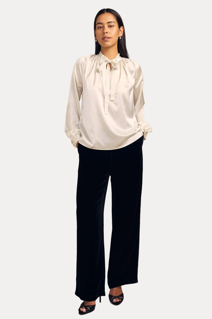 A woman modeling a white satin long-sleeve blouse with a tie-neck detail and black wide-leg velvet pants, standing against a solid beige studio background.