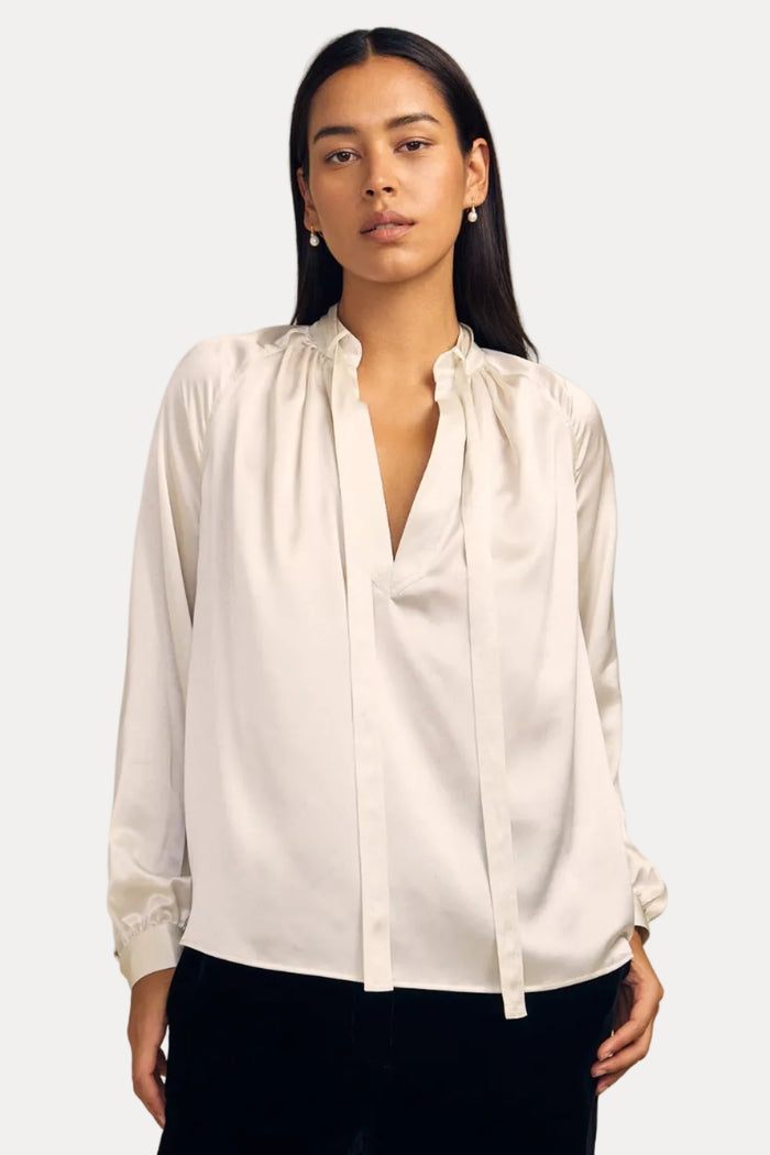 A woman modeling a white long-sleeve silk blouse with a gathered, tie-neck design, paired with black trousers, against a solid beige studio background.