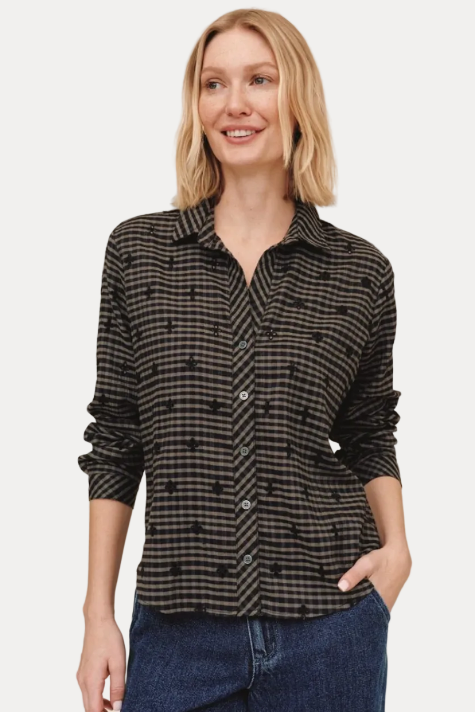 A woman modeling a black and gray patterned button-down shirt with dark blue jeans, standing against a solid beige background.