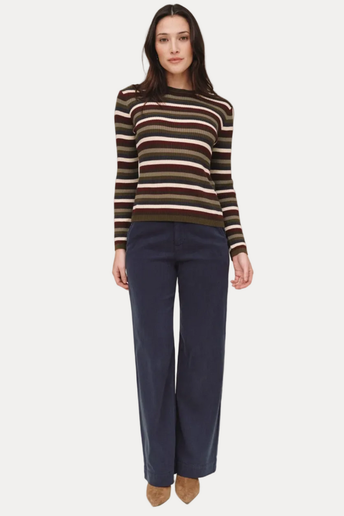 A woman stands against a plain beige studio background wearing a long-sleeve knit sweater with horizontal stripes in olive green, brown, cream, and black, paired with dark navy wide-leg pants and tan pointed shoes.