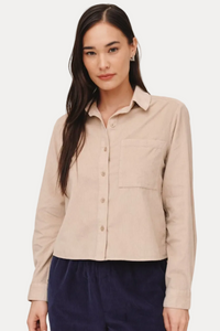 A woman modeling a light beige long-sleeve button-up shirt with a chest pocket, paired with dark navy pants, against a solid light beige background.