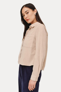 A woman wearing a light beige long-sleeve button-up shirt with a chest pocket and navy blue pants, photographed against a plain light beige studio background.