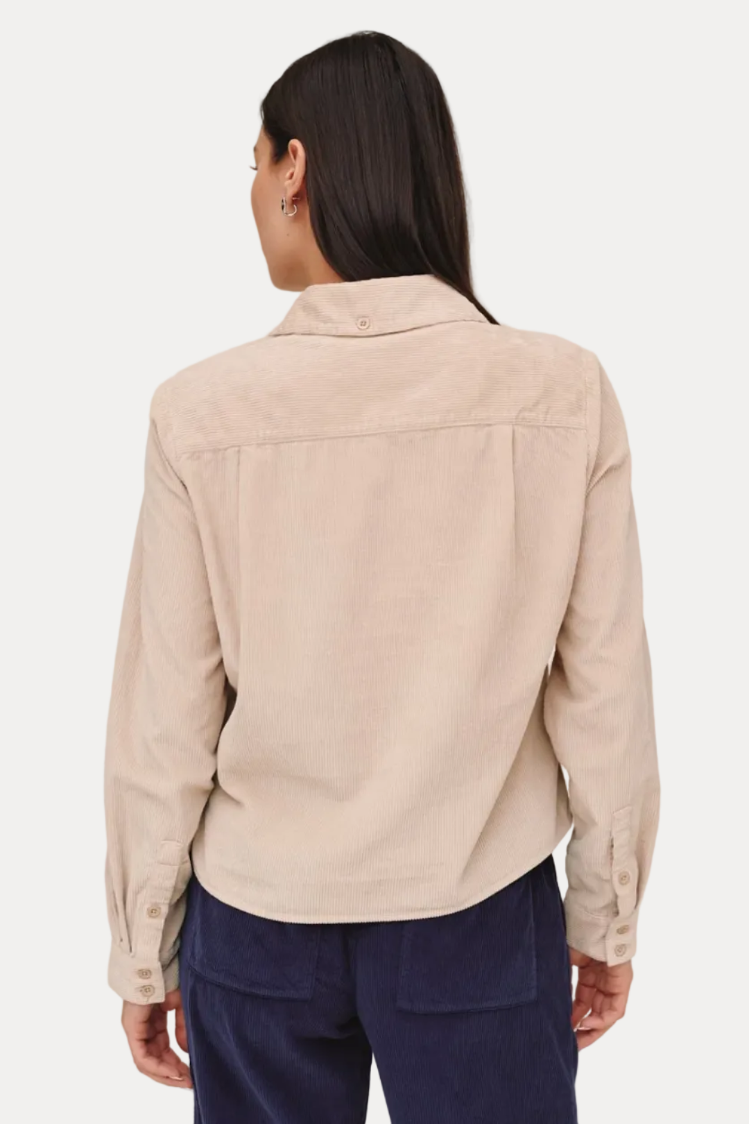 A woman shown from the back wearing a light beige corduroy shirt jacket and navy blue corduroy trousers, photographed against a solid light beige background.