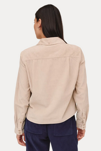 A woman shown from the back wearing a light beige corduroy shirt jacket and navy blue corduroy trousers, photographed against a solid light beige background.