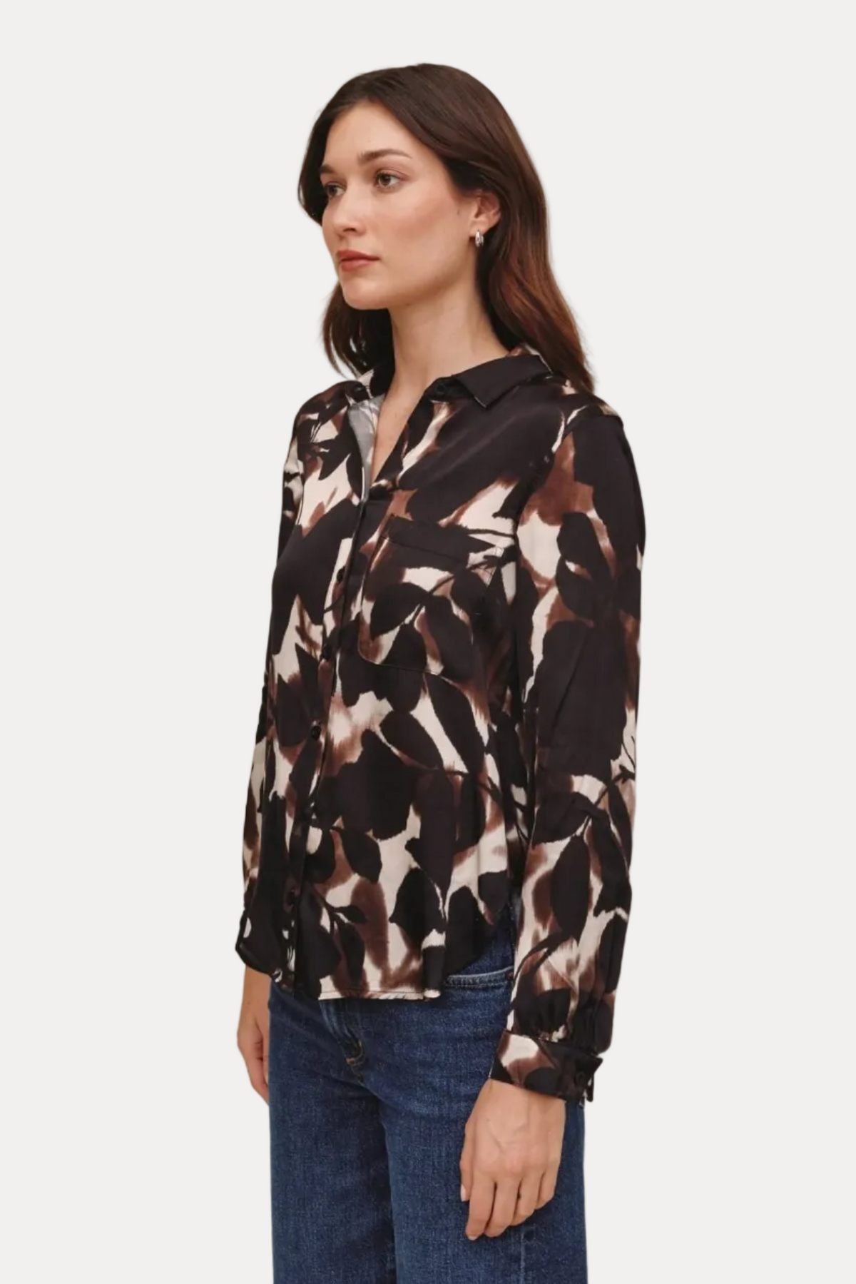A woman wearing a long-sleeve button-up blouse with an abstract brown, black, and beige pattern, paired with blue jeans, standing against a solid light beige background.