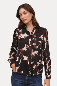 A woman modeling a long-sleeve button-up blouse with a dark brown, black, and beige abstract floral pattern, paired with blue jeans, against a solid light beige background.
