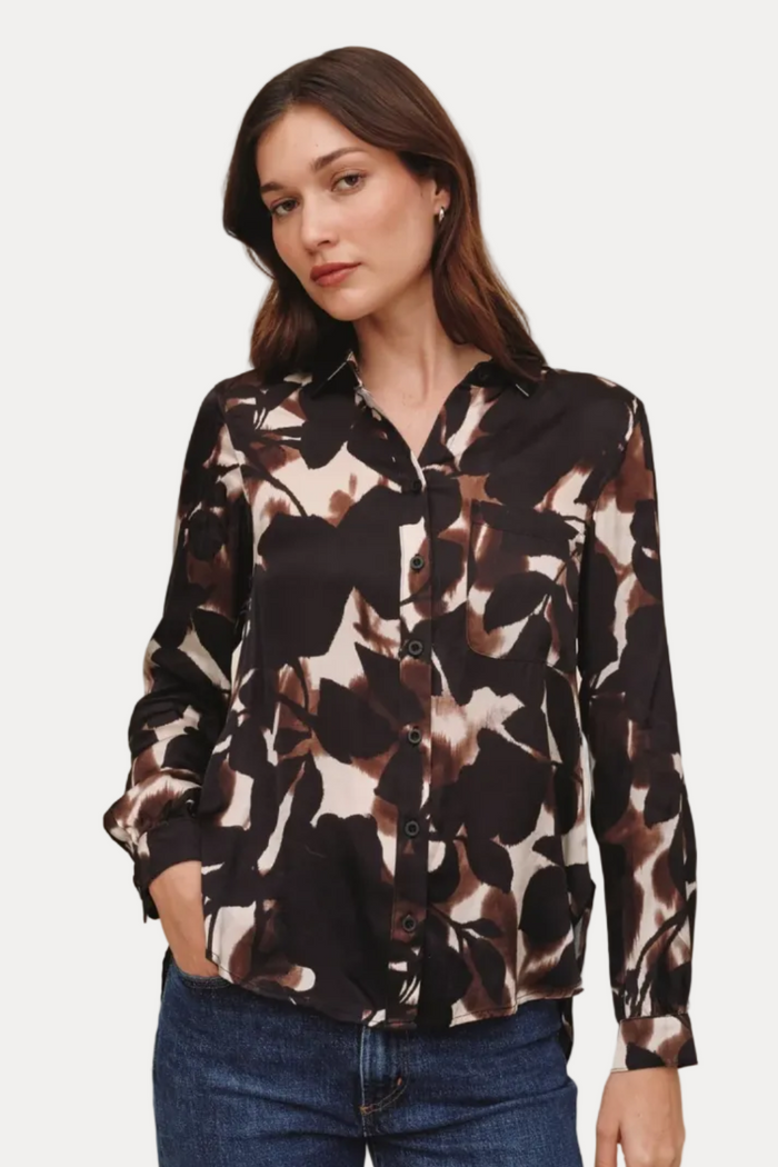 A woman modeling a long-sleeve button-up blouse with a dark brown, black, and beige abstract floral pattern, paired with blue jeans, against a solid light beige background.