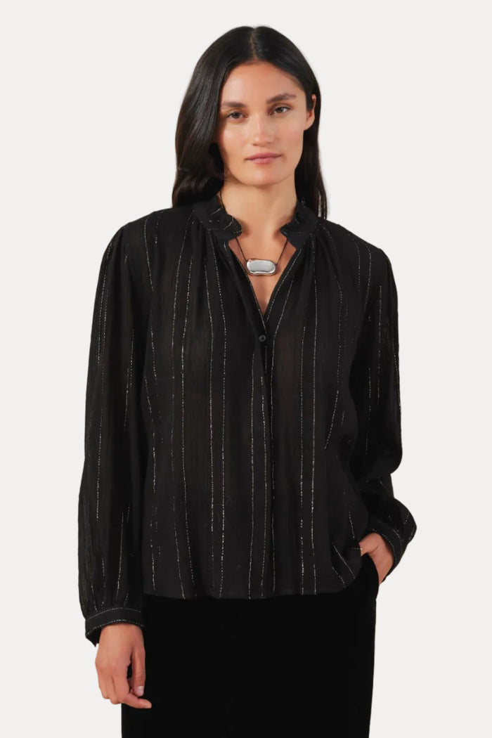 A woman modeling a black long-sleeve button-up blouse with thin vertical metallic-looking stripes, paired with black pants, against a plain light beige studio background.