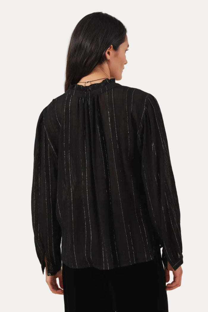 A woman shown from the back wearing a black long-sleeve blouse with subtle vertical metallic-looking stripes and black pants, photographed against a plain light beige studio background.
