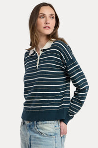 A woman modeling a navy blue and white horizontally striped long-sleeve polo sweater with a light beige collar, paired with light wash blue jeans, against a plain light gray studio background.