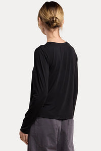 A woman shown from the back wearing a black long-sleeve crop top and dark gray wide-leg pants, photographed against a plain beige studio background.
