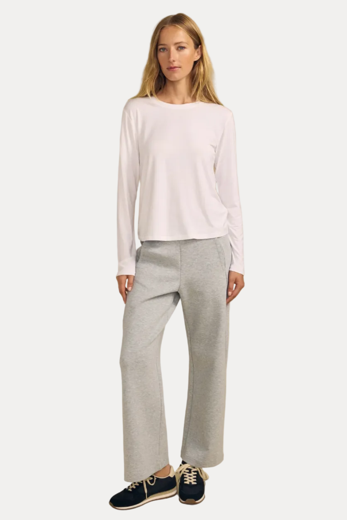 A woman modeling a white long-sleeve crewneck top and light gray wide-leg sweatpants, paired with navy blue sneakers, against a solid beige studio background.