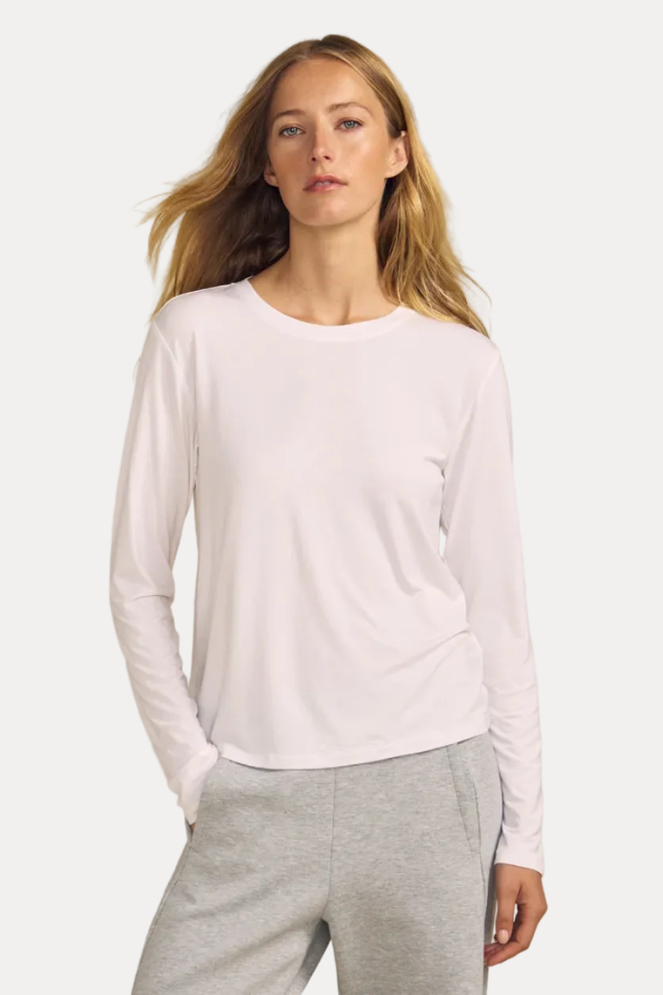 A woman modeling a white long-sleeve crewneck top and light gray heather sweatpants against a solid beige studio background.