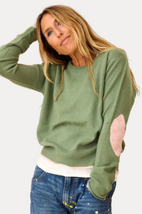 A woman wearing a sage green knit sweater with pink elbow patches, paired with blue jeans, standing in front of a colorful abstract painting.