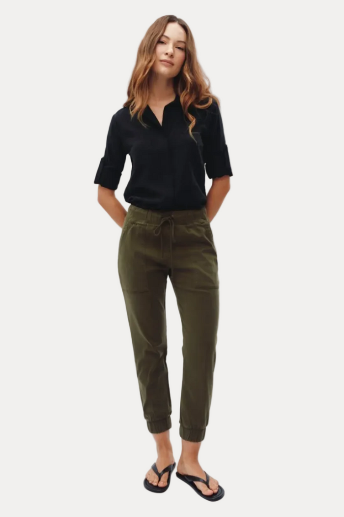 A woman modeling olive green drawstring cropped pants with a black shirt and black flip-flops, standing against a plain white studio background.