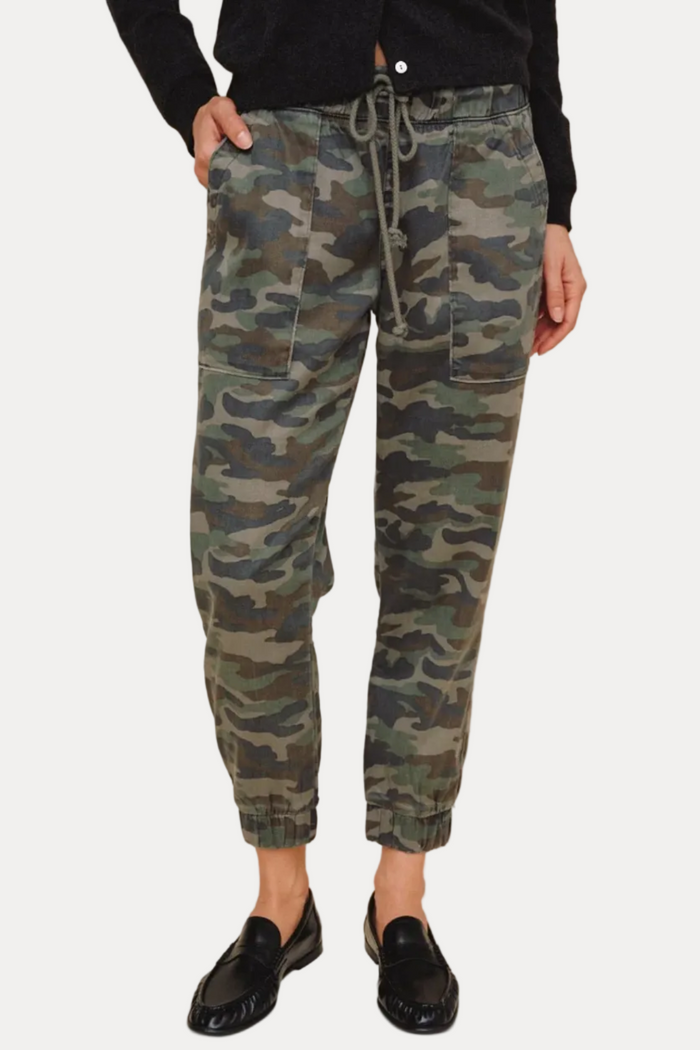 A person wearing camouflage jogger-style pants with a drawstring waist, shown from the waist down against a plain beige studio background. The pants have an allover camo pattern in green, brown, black, and beige tones, paired with black loafers.