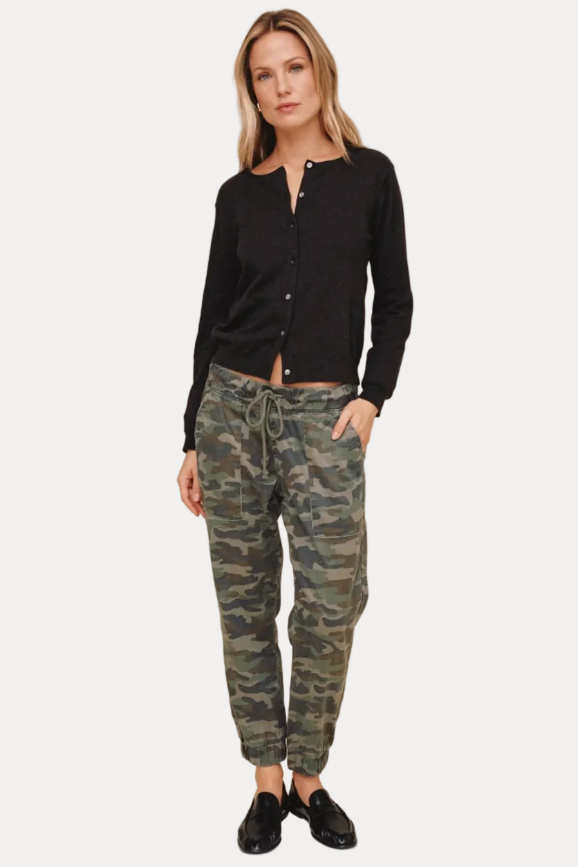 A woman modeling a black long-sleeve button-front cardigan and green camouflage jogger pants with black loafers, set against a solid beige studio background.