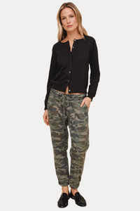 A woman modeling a black long-sleeve button-front cardigan and green camouflage jogger pants with black loafers, set against a solid beige studio background.