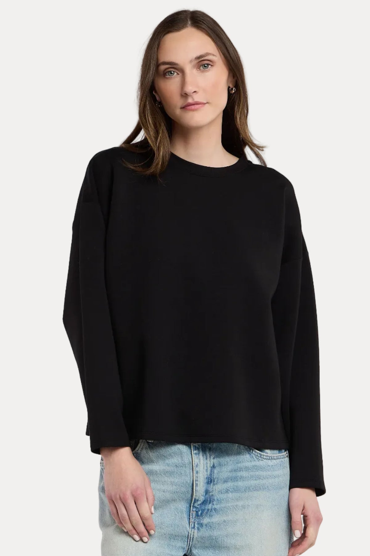 A woman wearing a black long-sleeve crewneck sweater and light wash blue jeans, standing against a plain light gray studio background.